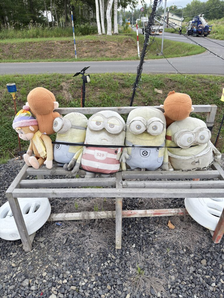 Faded Minions and Curious George plush toys lined up in front of Haga Farm’s unmanned farm stand in Hokkaido, creating a quirky and slightly eerie photo spot.