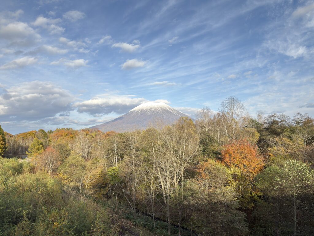 Mt. Yotei’s first snow and autumn foliage seen from Satsudora Bridge, Niseko, Hokkaido — a stunning view with blue skies, colorful forests, and the iconic snow-capped peak.